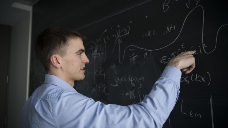 Man wearing a blue dress shirt working on equations at a chalkboard