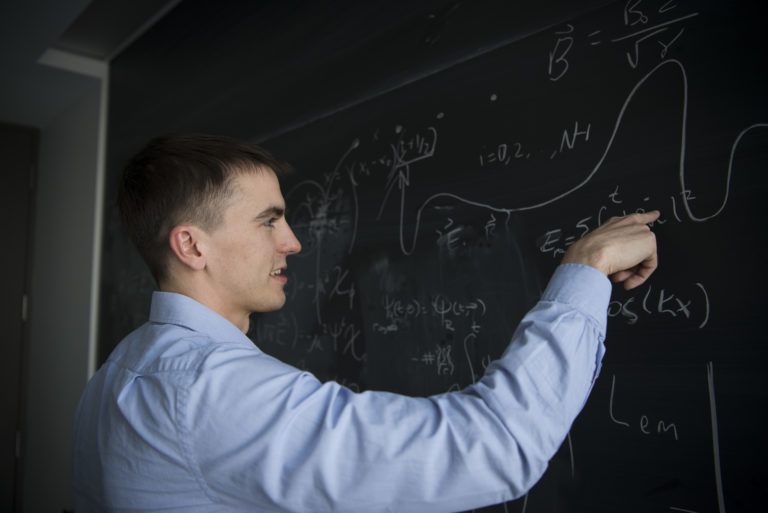 Man wearing a blue dress shirt working on equations at a chalkboard