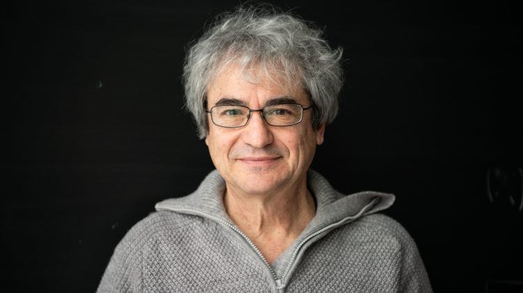 A portrait of a white man with grey hair and a grey sweater gazing warmly into the camera
