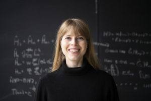 Associate Faculty member Christine Muschik. Photo of a blonde woman in a black sweater standing in front of a blackboard with equations on it.