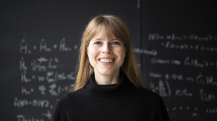 Associate Faculty member Christine Muschik. Photo of a blonde woman in a black sweater standing in front of a blackboard with equations on it.