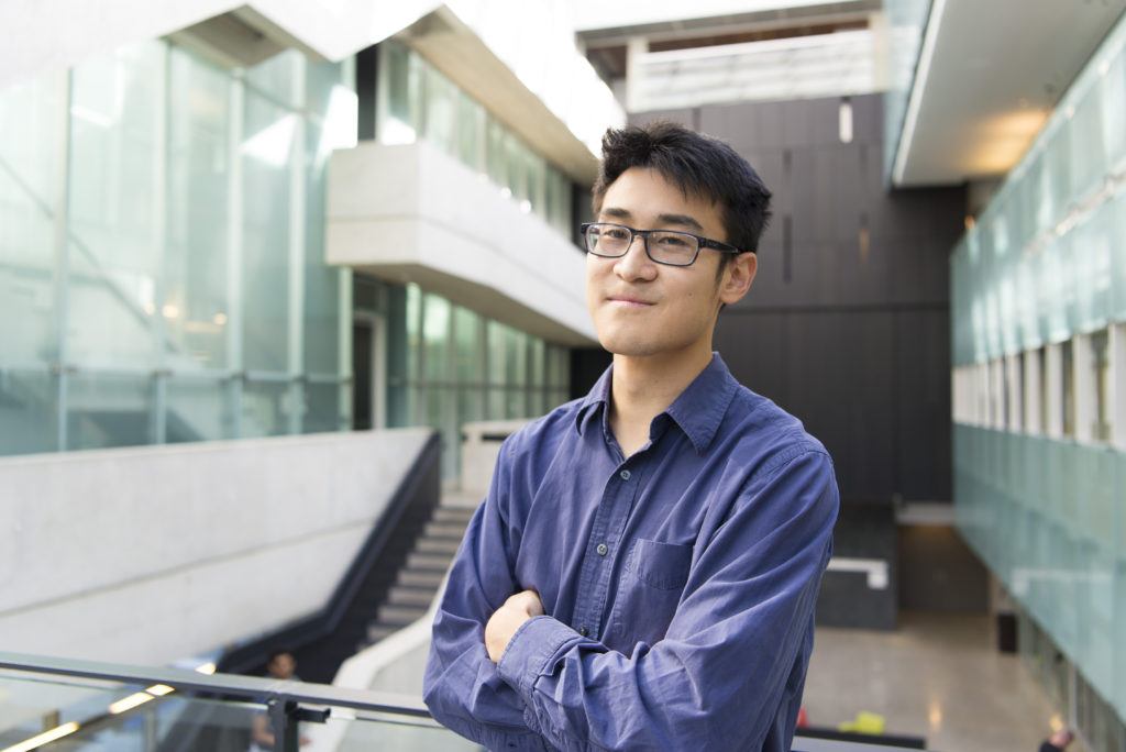 Photo of Tim Hsieh, a man with dark hair and glasses, wearing a blue shirt, standing in the atrium at Perimeter Institute
