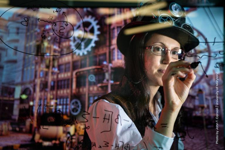 Researcher Nicole Yunger Halpern writes equations on a transparent board between her and the camera. She is wearing a top hat with gears on it.