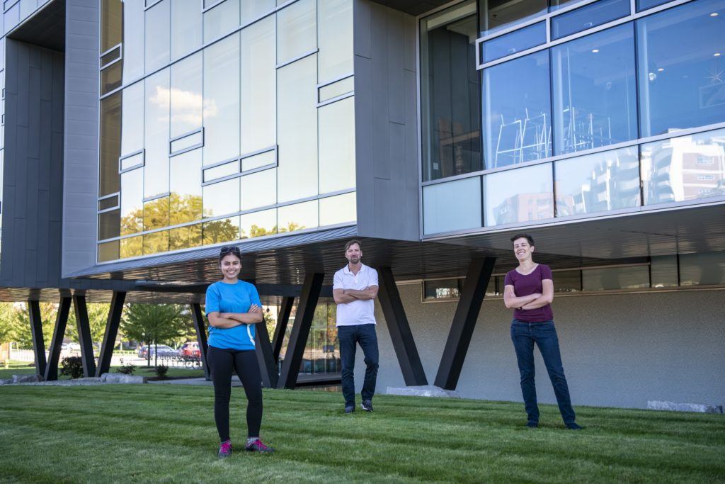 Three people stand on the grass outside the glass wall of the Perimeter Institute for Theoretical Physics