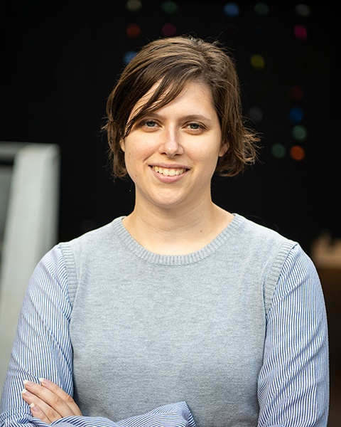 Portrait of a woman with short brown hair and grey and blue shirt standing in an atrium