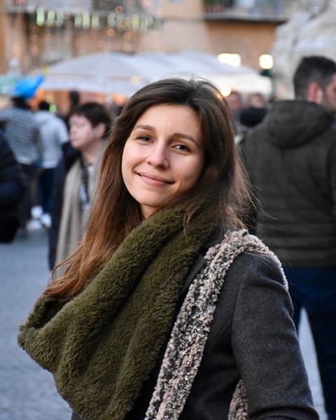 A woman with dark hair in a big scarf smiles in a crowded street.