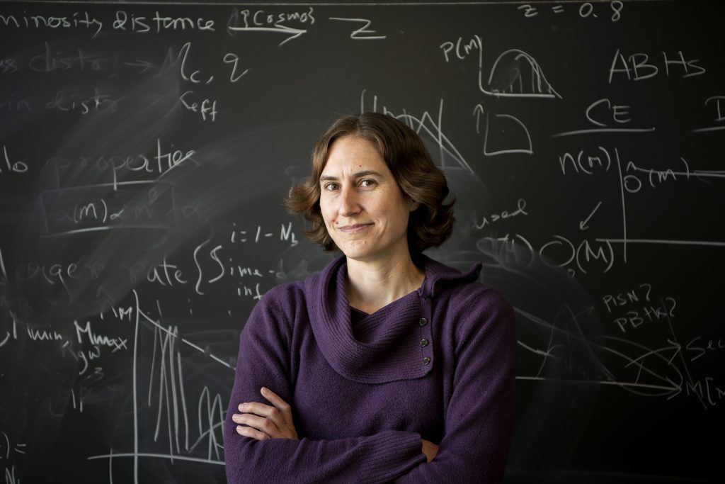 Woman with short brown hair wearing a purple sweater standing in front of a chalkboard of equations