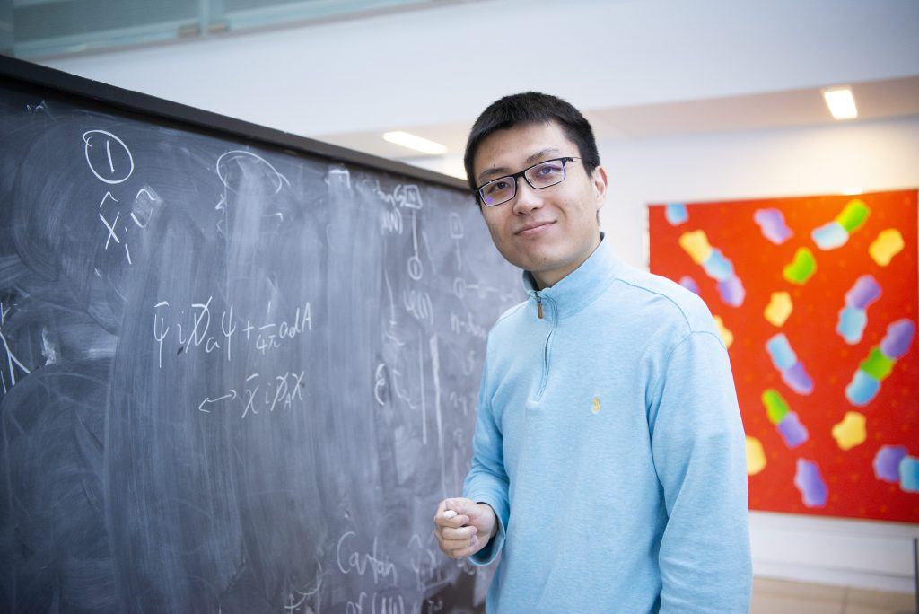 Homme portant des lunettes et un chandail bleu, debout devant un tableau noir où il a écrit des équations
