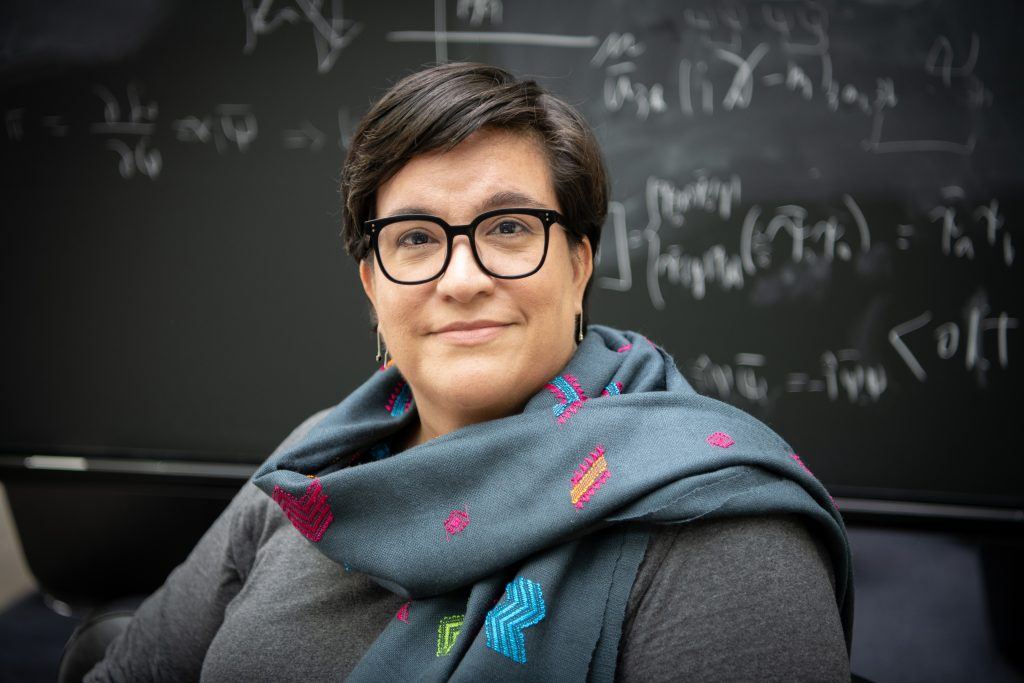 Woman in a grey shirt wearing a scarf and glasses siting in front of a chalkboard of equations